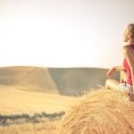 Woman with a backpack sitting on a hay bale, enjoying a serene sunset over a vast countryside field.